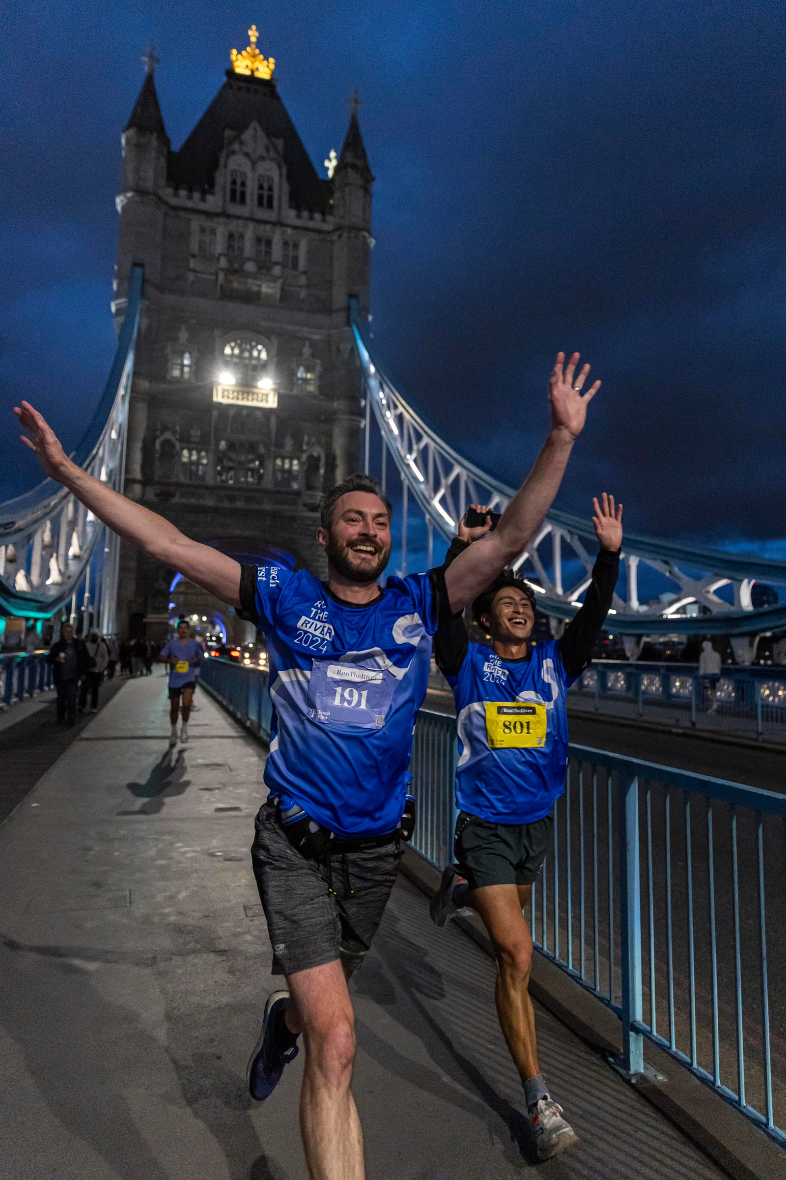 Two runners in blue jerseys sprint joyfully across Tower Bridge in London at dusk, arms raised in celebration, with the illuminated bridge and dark sky in the background.