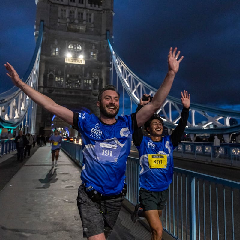 Two runners in blue jerseys sprint joyfully across Tower Bridge in London at dusk, arms raised in celebration, with the illuminated bridge and dark sky in the background.