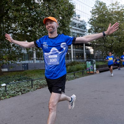 A smiling man in a blue running shirt and orange cap runs with arms outstretched during a race in a city park, with other runners and trees in the background.