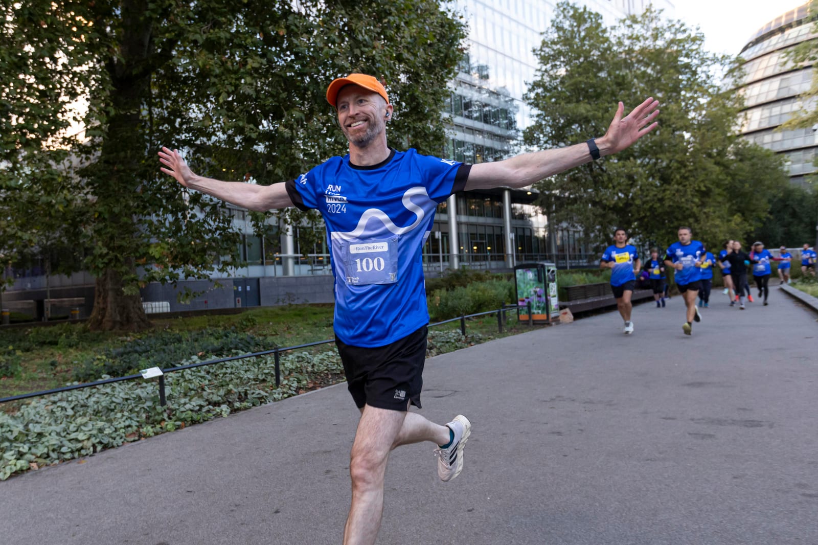 A smiling man in a blue running shirt and orange cap runs with arms outstretched during a race in a city park, with other runners and trees in the background.