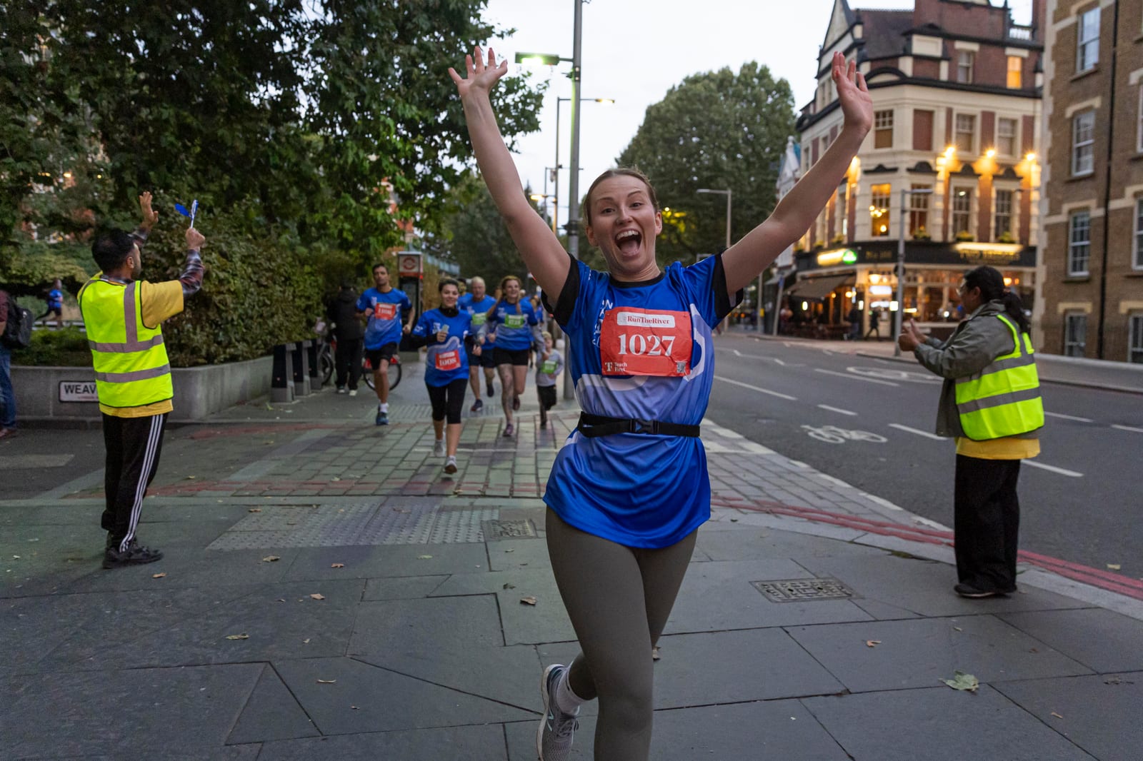 A smiling woman wearing a blue shirt and race bib 1027 raises her arms in celebration while running on a city street, with other runners and volunteers in yellow vests visible in the background.