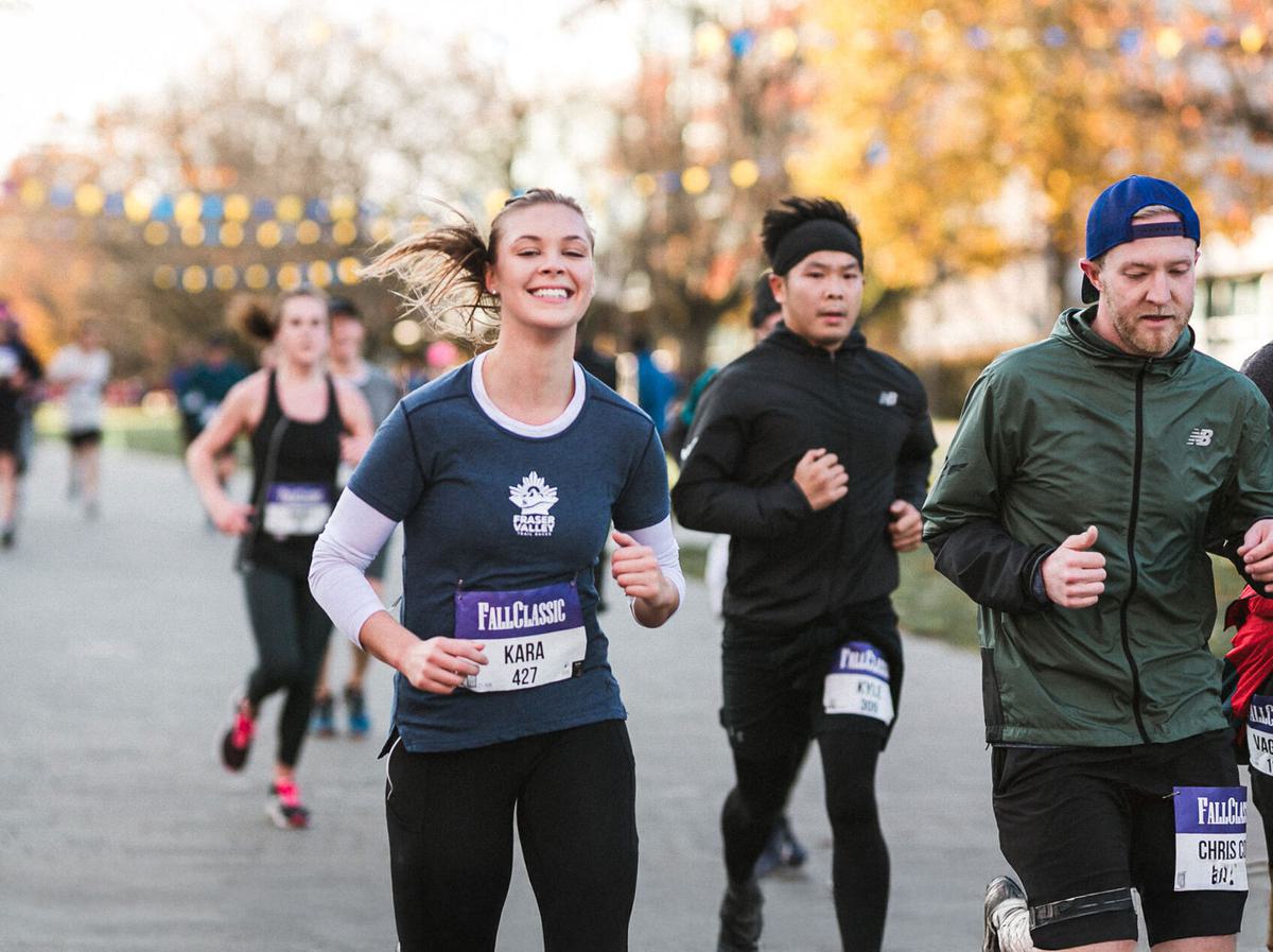 Group of people running in an outdoor race, with one smiling woman in the foreground wearing a blue shirt and race bib. Trees and other runners are visible in the background.