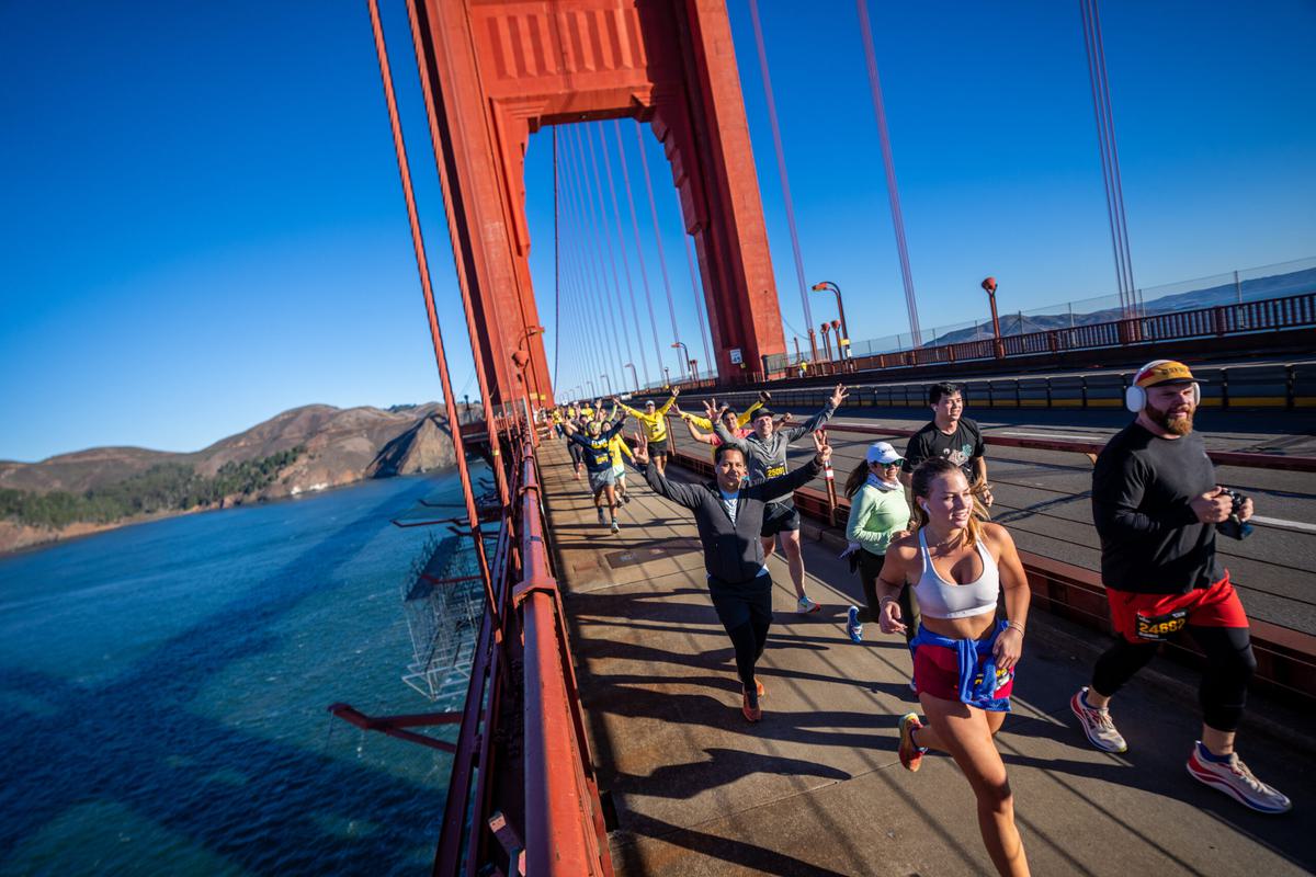 A group of marathon runners cross the Golden Gate Bridge on a sunny day, with blue sky overhead and water below. Some runners look joyful, raising their arms, while others focus ahead. Hills are visible in the background.