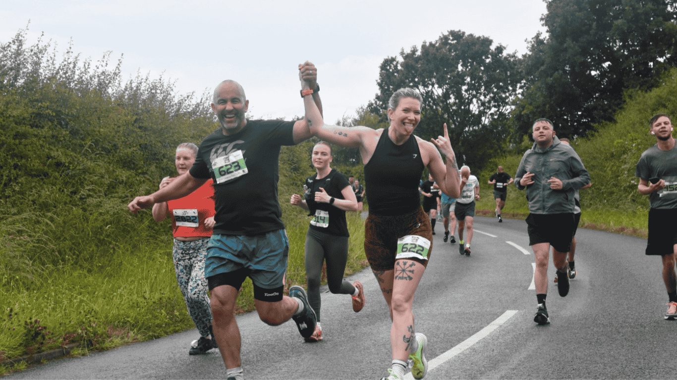 Runners participate in a race on a rural road. A man and woman hold hands, smiling and celebrating as they lead the group. Lush greenery and bushes line the road on both sides. Other runners follow behind, some focused and others cheerful.