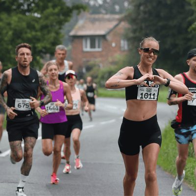 A group of runners participating in a race on a rural road, flanked by greenery. One woman in the foreground is smiling and gesturing with her hands. Participants wear numbered bibs and a variety of athletic gear.