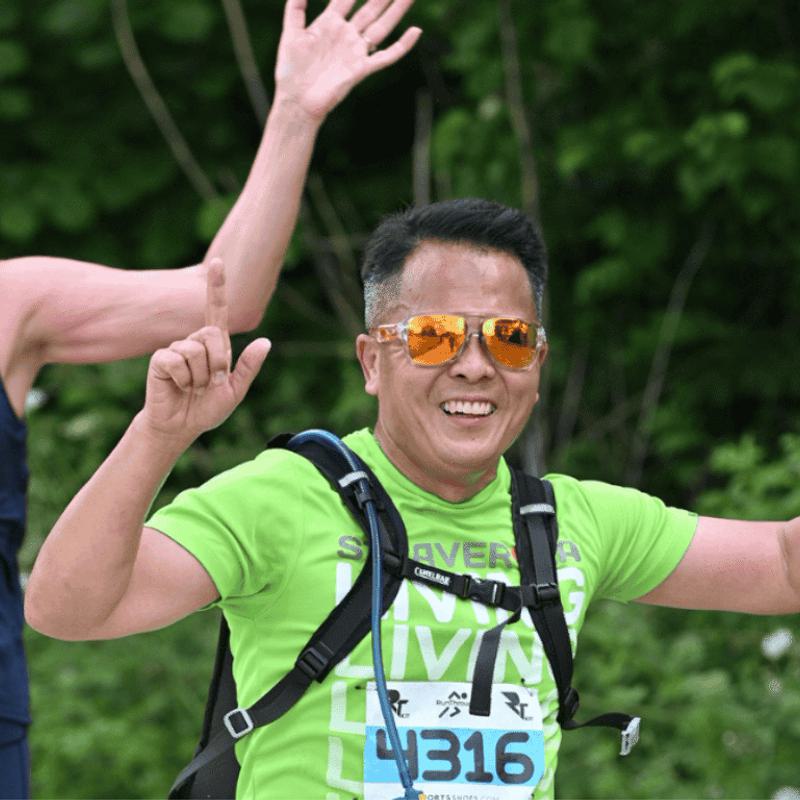 A man and a woman are running outdoors, smiling and waving. The man is wearing orange reflective sunglasses and a green shirt with the number 4316. The woman is wearing a navy tank top with the number 4328. Green foliage is in the background.