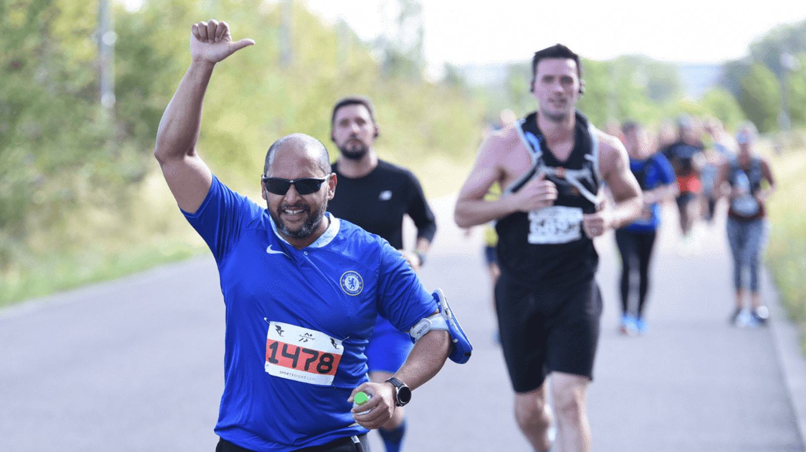 A man in a blue sports shirt runs and raises his hand in a thumbs-up gesture during a race. Other runners are visible in the background on a paved path lined with greenery.