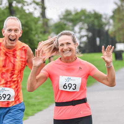 Two runners, a man in an orange sleeveless top and a woman in a coral top, smile and wave at the camera during a race. Both wear bib numbers, with a park and other runners visible in the background.
