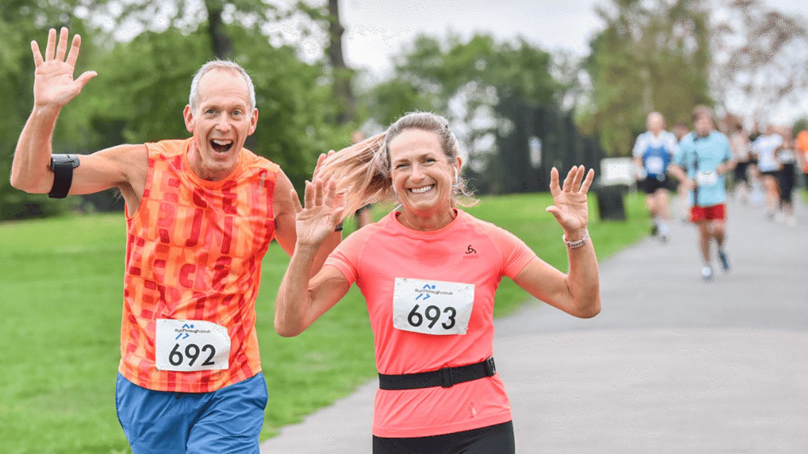 Two runners, a man in an orange sleeveless top and a woman in a coral top, smile and wave at the camera during a race. Both wear bib numbers, with a park and other runners visible in the background.