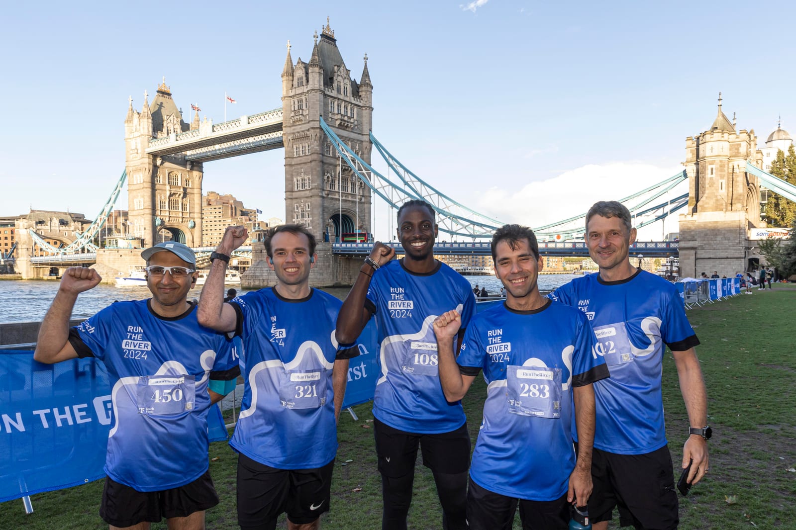 Five runners in matching blue shirts stand smiling with raised fists in front of Tower Bridge in London, celebrating after a race on a sunny day.