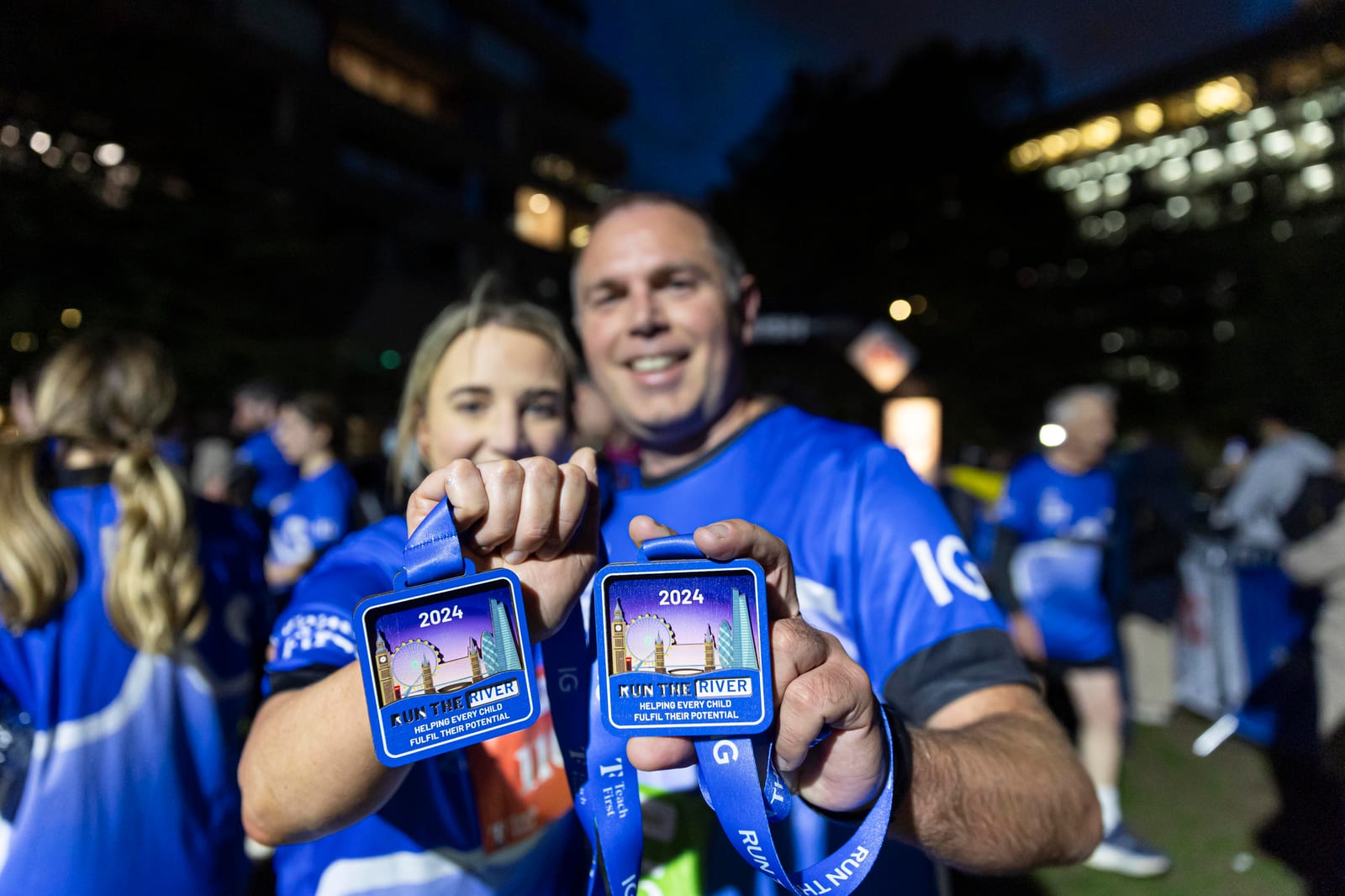 Two runners proudly display finisher medals for the 2024 “Run the River” event, smiling at the camera. Both wear blue race shirts, with other participants visible in the background at night.