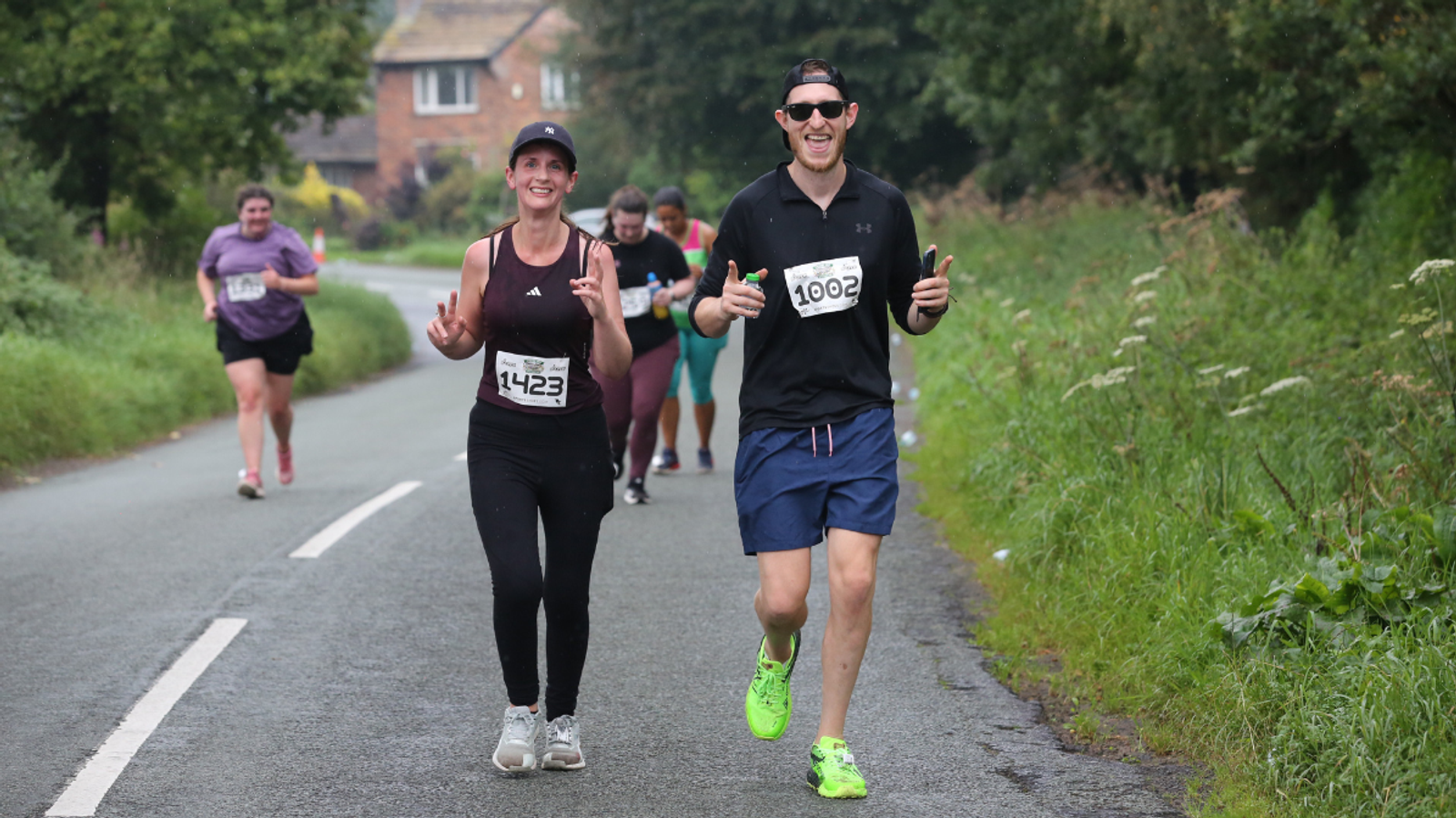 Two runners in athletic gear smile and gesture with their hands while jogging on a rural road, surrounded by greenery and other participants in the background. Both are wearing numbered bibs, indicating they are in a race.